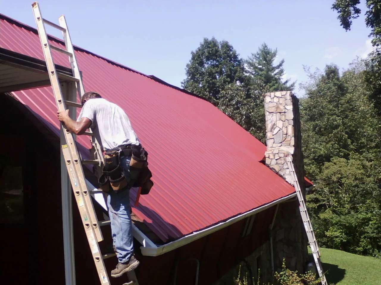 Roofer on a ladder beside a red metal roof and stone chimney — Soffit & Fascia Repair by Roofing Brothers in Rixeyville